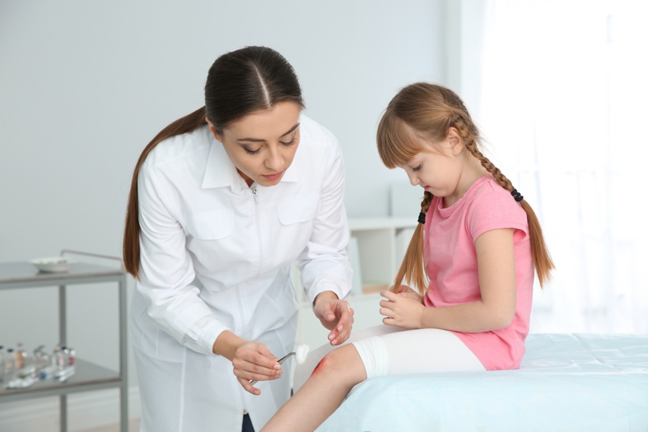 Female doctor cleaning little girl's leg injury in clinic. First aid