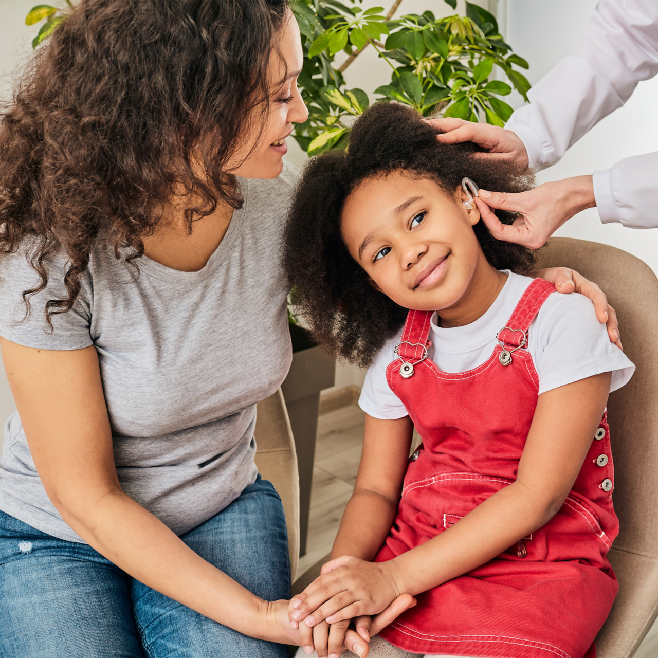 A young girl, smiling, holds her mother's hand while receiving a hearing aid. 