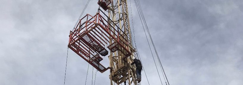 A man in dark clothing climbs down an oil rig against a cloudy sky with state office buildings in the background