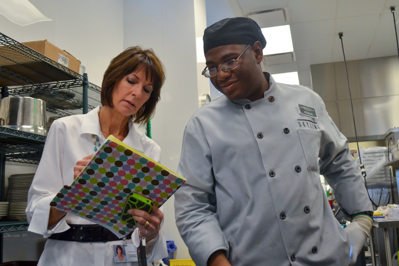 A woman with a notebook goes over something with a young man in kitchen workers uniform.