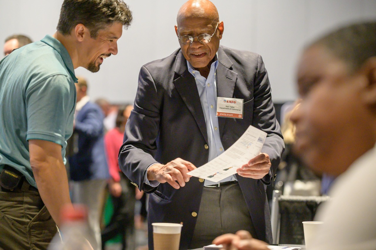 Department of Commerce employee Ken Talley showing a flyer to a smiling attendee.