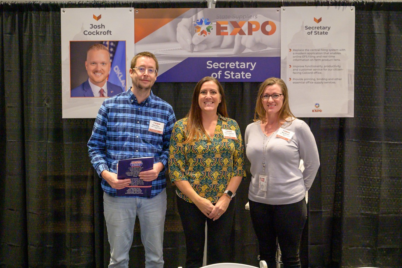 Three cabinet staff smile in front of the Secretary of State booth.