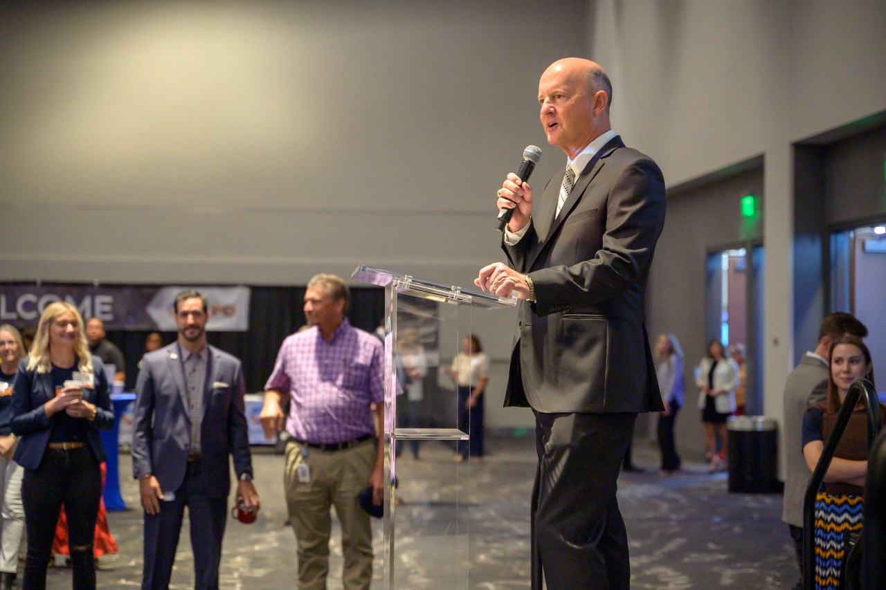 State Chief Operating Officer John Suter speaks with microphone on ballroom stage.